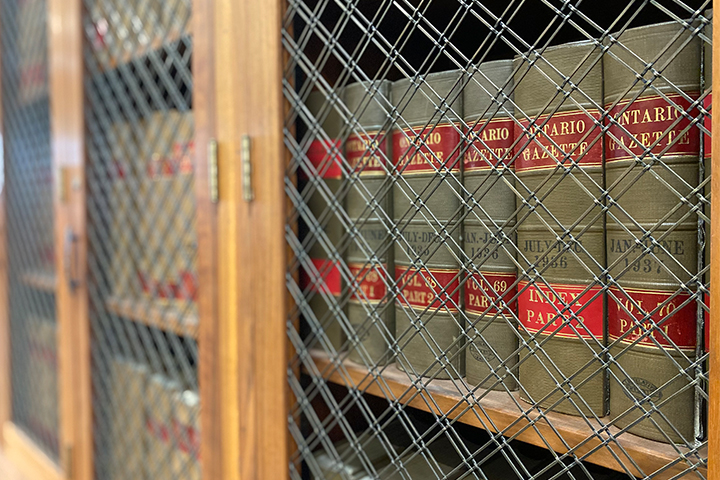 Des exemplaires de la Gazette de l’Ontario alignés derrière le grillage métallique d’armoire en bois.